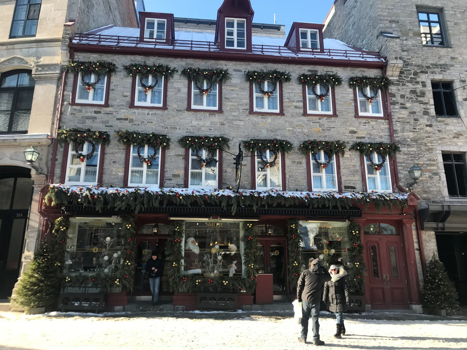 A stone shopfront in Old Québec dressed for Christmas with wreaths and garlands, snow banked on the street.